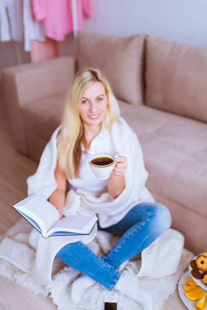 Beautiful smiling blonde female sits beside the sofa, keeps a book in right hand and cup of coffee in left hand at home - Фото, изображение