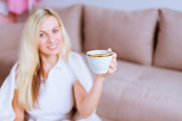 Beautiful pretty woman sits beside the couch and keeps cup of coffee in her hand in the foreground - 写真・画像