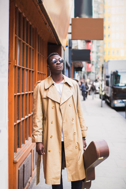 Portrait of young handsome stylish man who stands with guitar case in hand on sidewalk of the street - Fotó, kép