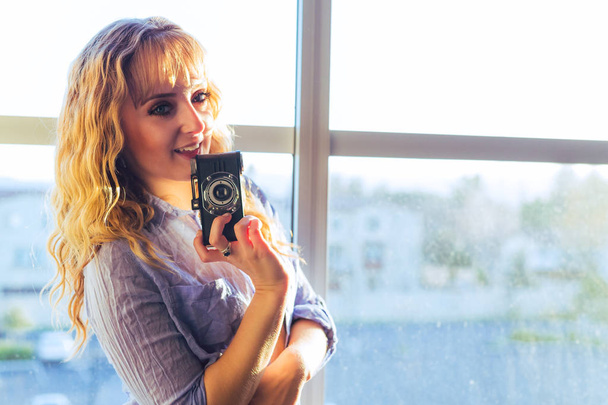 Νεαρή γυναίκα Καυκάσιος attactive με μεγάλα μάτια και ξεκούμπωτο γαλάζιο πουκάμισο ποζάρει με ρετρό κάμερα από το παράθυρο με χρυσή ώρα φως - Φωτογραφία, εικόνα