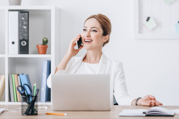 feliz exitosa mujer de negocios hablando en el teléfono inteligente en la oficina moderna con el ordenador portátil
 - Foto, Imagen