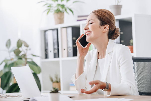 смеющаяся деловая женщина разговаривает на смартфоне в офисе с ноутбуком
 - Фото, изображение