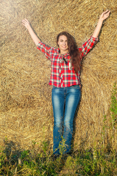 Country style concept. Young beautiful woman in jeans and a plaid shirt stands near a haystack with arms raised in front of her and smiling in the rays of the sunset - Photo, Image