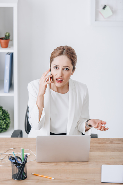 confused emotional businesswoman talking on smartphone at workplace with laptop - Foto, Bild