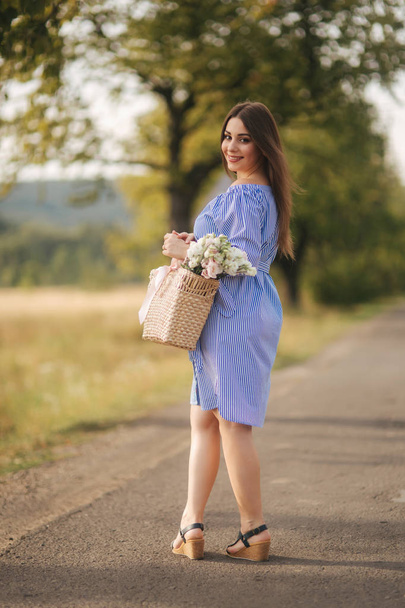 Красива вагітна жінка в блакитній сукні, що йде, чує ферму. В'язана сумка з букетом квітів. Фон дерева і гір
 - Фото, зображення