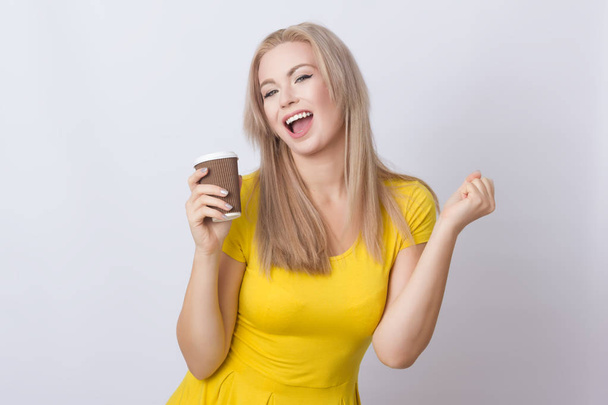 Cute blonde woman in yellow dress holding coffee cup in her hands. over grey backgroun - Fotografie, Obrázek