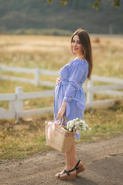 Beautiful pregnant woman in blue dress walking hear the farm. Knitted bag with bouquet of flowers. Background of tree and mountains - Fotó, kép