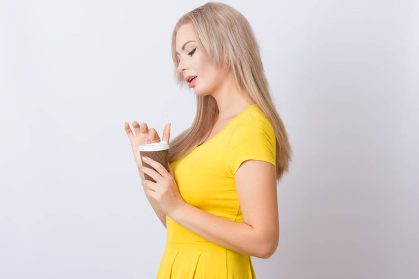 Cute blonde woman in yellow dress holding coffee cup in her hands. over grey backgroun - Φωτογραφία, εικόνα