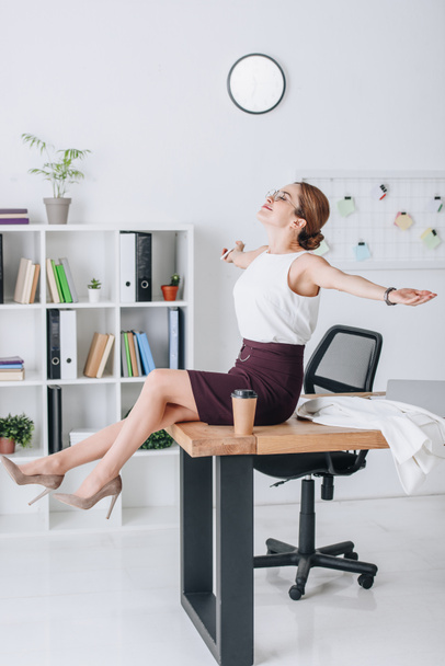 happy businesswoman with outstretched hands sitting on table during coffee break in modern office - Фото, изображение