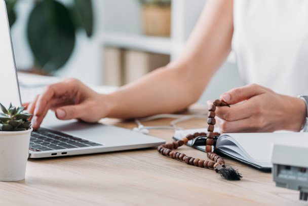 abgeschnittene Ansicht einer Geschäftsfrau mit Laptop und Rosenkranzperlen am Arbeitsplatz - Foto, Bild