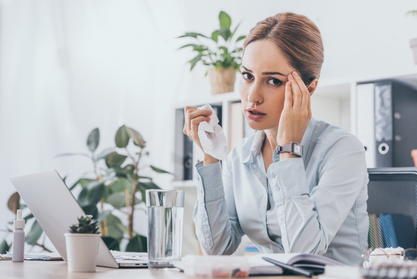 sick adult businesswoman sitting at workplace with glass of water and looking at camera while touching her head - Photo, Image