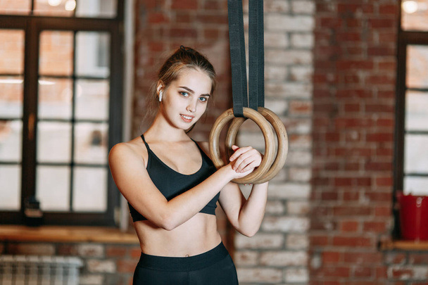 Sports girl doing stretching in the gym with rings Stands and smiles, holding on to the rings, rest in training. Professional equipment and clothing - Фото, зображення