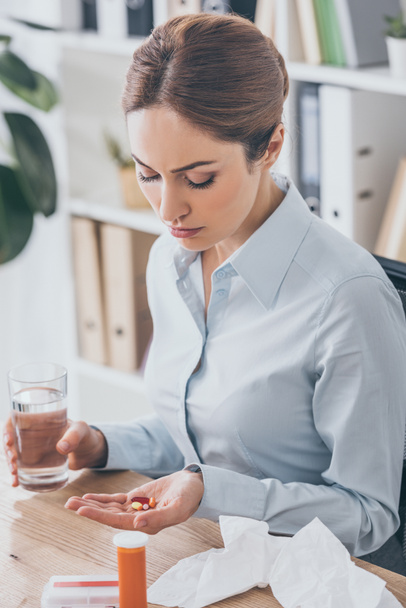 diseased businesswoman sitting at workplace with pills and glass of water - Photo, image