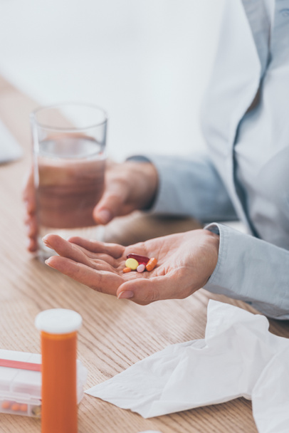 cropped shot of sick businesswoman sitting at workplace with pills and glass of water - Foto, Imagen