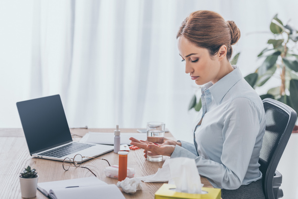 sick adult businesswoman sitting at workplace with pills and glass of water - Photo, Image