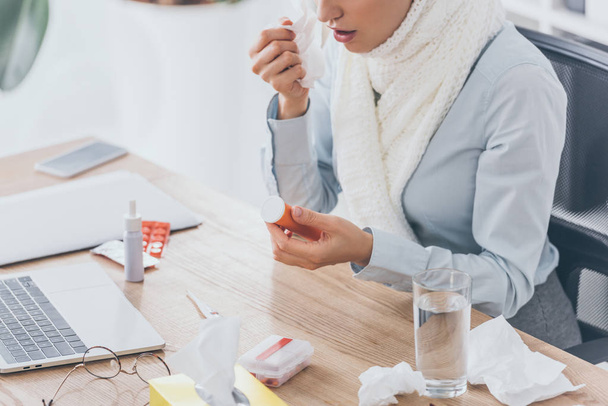 cropped shot of sick businesswoman in scarf holding container of pills at workplace - Valokuva, kuva