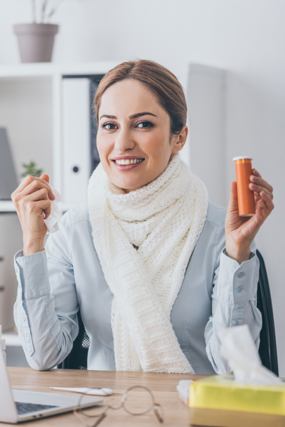 sick smiling businesswoman in scarf holding container of pills at workplace and looking at camera - Photo, Image