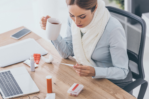 high angle view of sick businesswoman in scarf holding cup of tea and electric thermometer sitting at office - Photo, Image