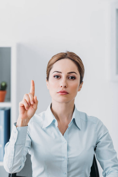 serious adult businesswoman looking at camera and pointing up at office - Foto, afbeelding