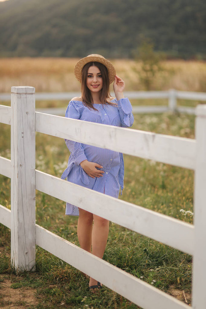 Beautiful pregnant woman in blue dress and knitted hat stand near the fance in the farm. She put hands on her belly and smile - Fotografie, Obrázek