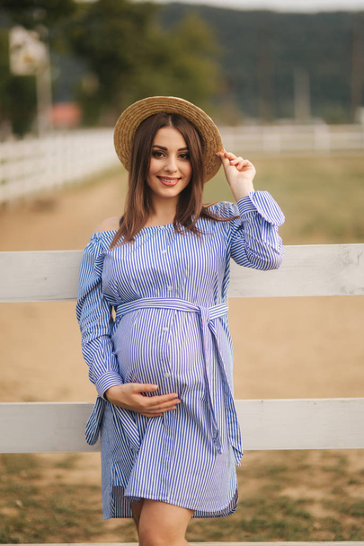 Beautiful pregnant woman in blue dress and knitted hat stand near the fance in the farm. She put hands on her belly and on her hat - Фото, изображение