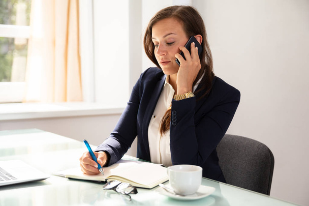 Бізнес-леді розмовляє на мобільному телефоні під час написання розкладу в щоденнику
 - Фото, зображення