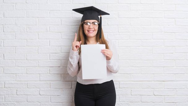 Молода доросла жінка над білою цегляною стіною в шапці випускника, що тримає ступінь здивований ідеєю або питанням, що вказує пальцем щасливе обличчя, номер один
 - Фото, зображення