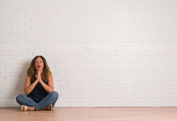 Hispanische Frau mittleren Alters sitzt auf dem Boden über weißen Ziegelsteinen und bettelt und betet mit Händen und hoffnungsvollem Gesichtsausdruck sehr emotional und besorgt. Bitte um Vergebung. Religionskonzept. - Foto, Bild