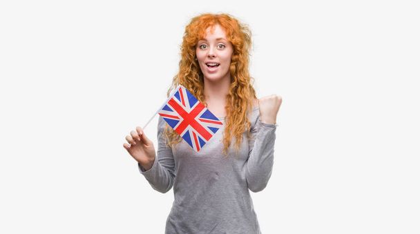 Joven pelirroja sosteniendo bandera del Reino Unido gritando orgullosa y celebrando la victoria y el éxito muy emocionada, animando emoción
 - Foto, Imagen