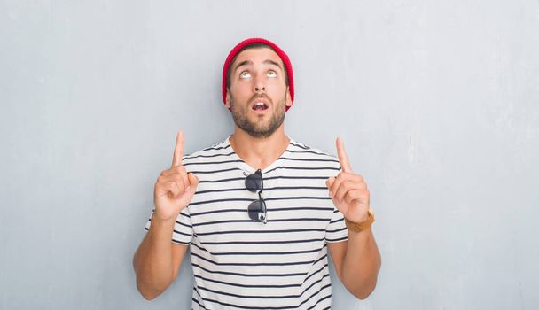 Beau jeune homme hipster sur le mur gris grunge portant t-shirt marine et bonnet de laine étonné et surpris de regarder vers le haut et pointant avec les doigts et les bras levés
. - Photo, image