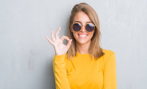 Красива молода жінка стоїть над гранжевою сірою стіною в ретро сонцезахисних окулярах робить знак з пальцями, відмінний символ
 - Фото, зображення