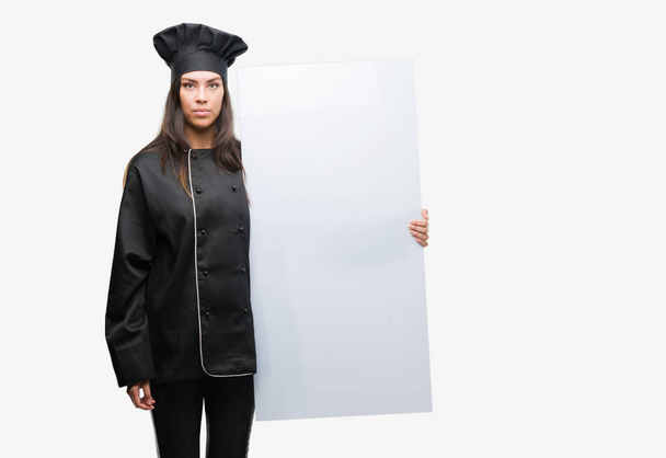 Young hispanic cook woman wearing chef uniform holding banner with a confident expression on smart face thinking serious - Photo, Image
