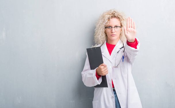 junge blonde Ärztin über grunge-graue Wand, Klemmbrett mit offener Hand haltend, Stoppschild mit ernstem und selbstsicherem Gesichtsausdruck, Verteidigungsgeste - Foto, Bild