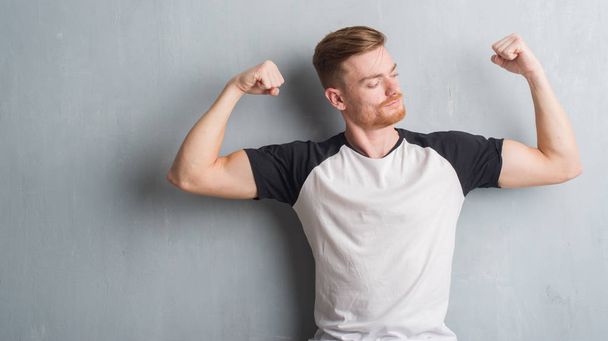 Jonge roodharige man over grijs grunge muur armen spieren glimlachend trots tonen. Fitness concept. - Foto, afbeelding
