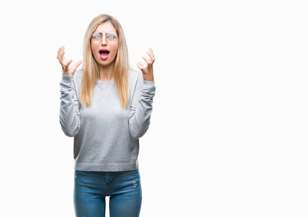 Young beautiful blonde woman wearing glasses over isolated background crazy and mad shouting and yelling with aggressive expression and arms raised. Frustration concept. - 写真・画像