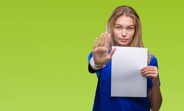 Young caucasian woman holding blank paper sheet over isolated background with open hand doing stop sign with serious and confident expression, defense gesture - Photo, Image