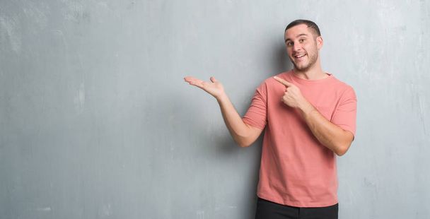 Молодий кавказький чоловік над сірою гранжевою стіною здивований і посміхається камері, представляючи рукою і вказуючи пальцем
. - Фото, зображення