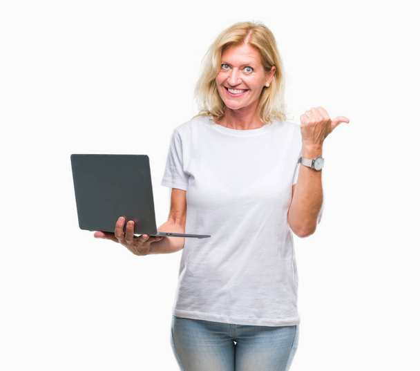Блондинка средних лет с помощью компьютера ноутбук на изолированном фоне указывая и показывая с большим пальцем в сторону со счастливым лицом улыбаясь
 - Фото, изображение