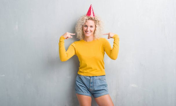 Νεαρή ξανθιά γυναίκα πάνω από το grunge γκρίζο τοίχο φορώντας καπέλο γενεθλίων αναζητούν αυτοπεποίθηση με χαμόγελο στο πρόσωπο, δείχνοντας τον εαυτό του με τα δάχτυλα, περήφανος και χαρούμενος. - Φωτογραφία, εικόνα