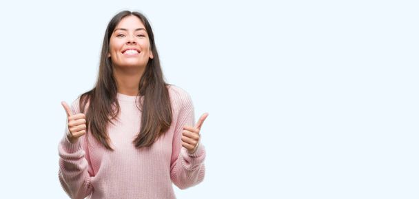 Young beautiful hispanic woman wearing a sweater success sign doing positive gesture with hand, thumbs up smiling and happy. Looking at the camera with cheerful expression, winner gesture. - Foto, Imagem