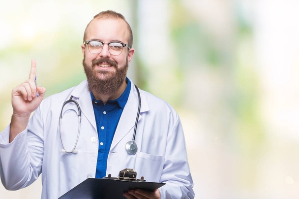 Young doctor man holding clipboard over isolated background surprised with an idea or question pointing finger with happy face, number one - Photo, Image