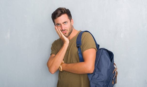 Handsome tourist young man over grey grunge wall wearing backpack thinking looking tired and bored with depression problems with crossed arms. - Fotografie, Obrázek