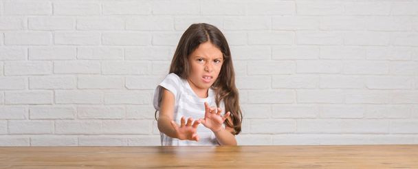 Nuori latino lapsi istuu pöydällä kotona iljettävä ilme, tyytymätön ja pelokas tekee inhoa kasvot, koska vastenmielisyys reaktio. Kädet ylhäällä. Ärsyttävä käsite
. - Valokuva, kuva
