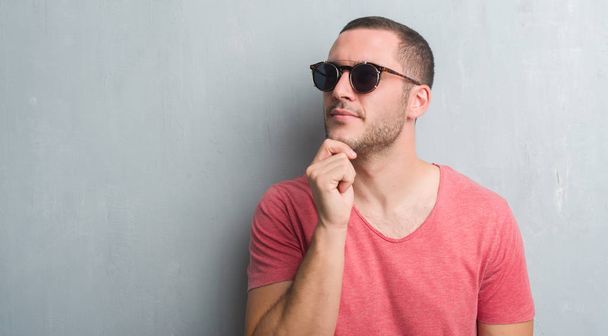 Kaukázusi fiatalember visel napszemüveg súlyos arc gondolkodás kérdése, nagyon zavaros ötlet szürke grunge falon - Fotó, kép