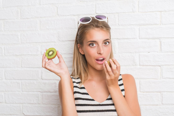 Beautiful young woman over white brick wall eating green kiwi with a confident expression on smart face thinking serious - Photo, Image