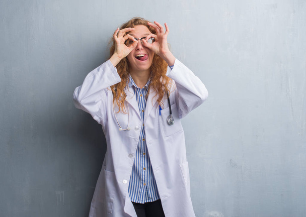 Joven doctora pelirroja sobre una pared de grunge gris usando un abrigo haciendo un gesto aceptable como binoculares sacando la lengua, ojos mirando a través de los dedos. Expresión loca
. - Foto, imagen