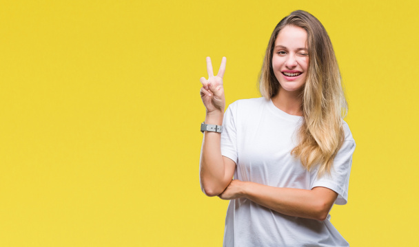 Young beautiful blonde woman wearing casual white t-shirt over isolated background smiling with happy face winking at the camera doing victory sign. Number two. - Photo, Image