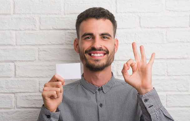 Jeune homme adulte sur le mur de briques tenant la carte blanche faisant signe ok avec les doigts, excellent symbole
 - Photo, image