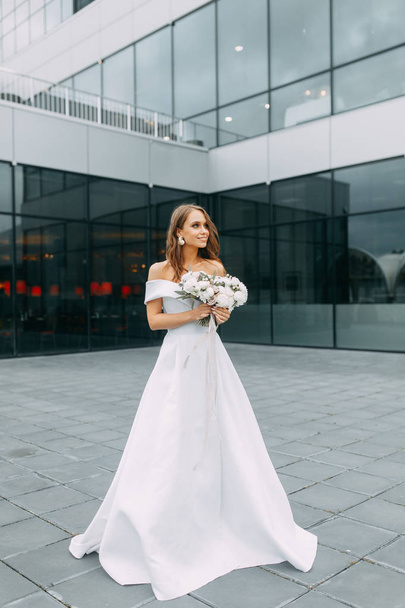 Modern, stylish bride in European dress. Photo shoot on the street near the hotel. Bouquet and strict dress - Foto, afbeelding