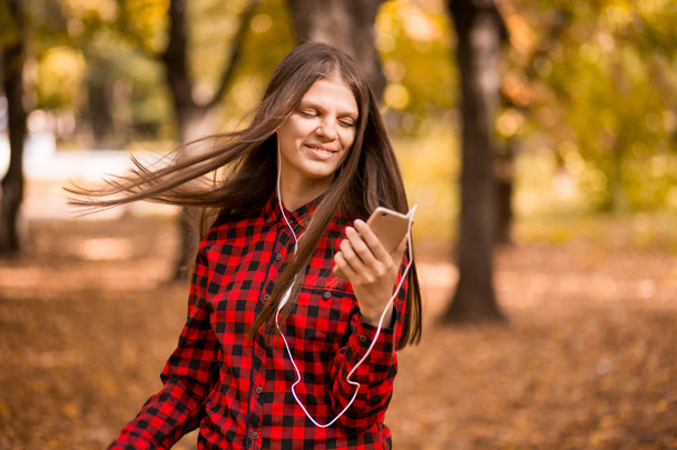 Pretty girl holding phone and listening music with her headphones in autumn park - Foto, imagen
